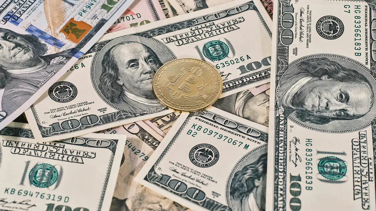 A physical bitcoin coin resting on a spread of US one hundred dollar bills, representing cash-to-crypto transactions